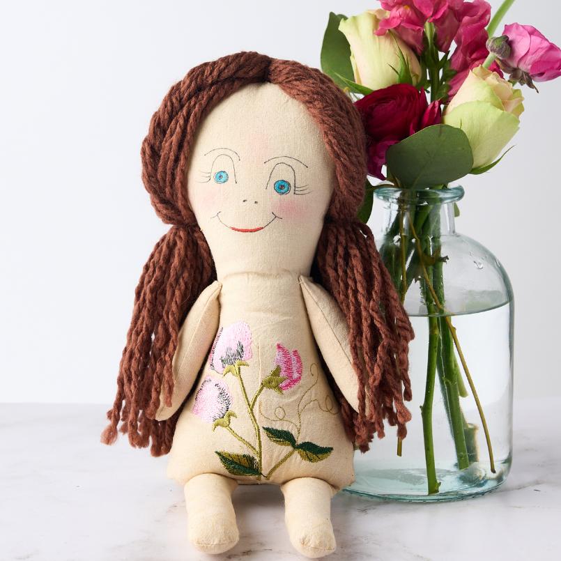 Close-up of the Sweet Pea Elegance Doll with intricate floral embroidery, placed beside a glass vase of flowers.