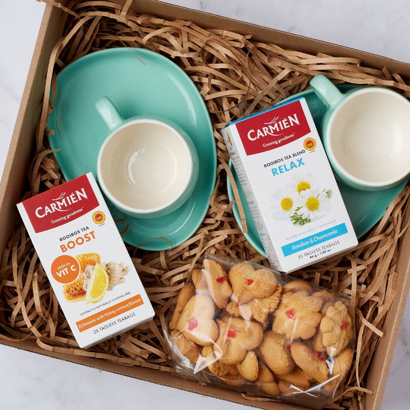 Close-up of gift box with Carmién Rooibos tea boxes in Boost and Relax blends, teal teacups, and heart-shaped biscuits.