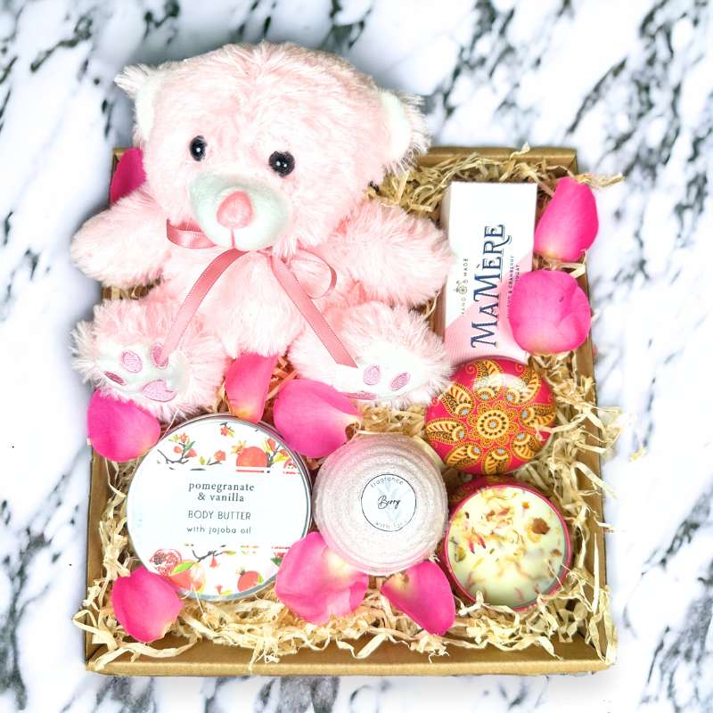 Pink teddy bear gift box with body butter, chocolate, bath bomb, and candle, surrounded by rose petals. Perfect for pampering and care.