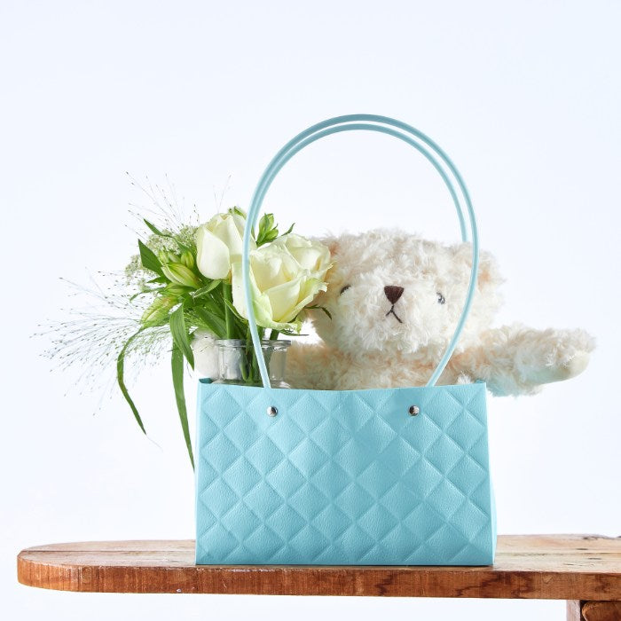 Teddy's Blossom Basket featuring a plush teddy bear peeking out of an aqua blue quilted basket with fresh white roses and green foliage, perfect for a unique baby shower gift.