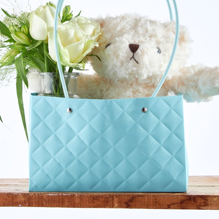 Close-up of Teddy's Blossom Basket with a soft teddy bear surrounded by delicate white roses in an elegant aqua blue tote, ideal for a luxury baby gift.