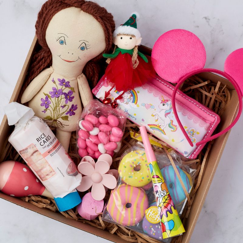 Twinkle Treats and Tidings gift box featuring a handcrafted doll, festive elf figurine, unicorn pencil case, headband, pink candy, rice cakes, donut bath bombs, flower soap, and a party blower.