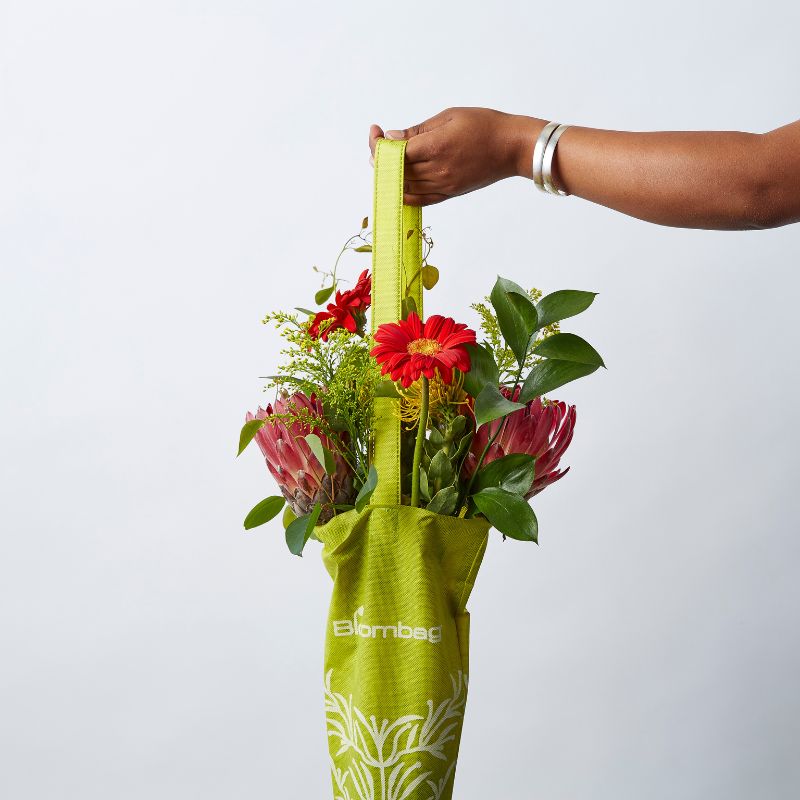 Hand holding a green reusable Bloombag tote filled with vibrant red and pink flowers and greenery.