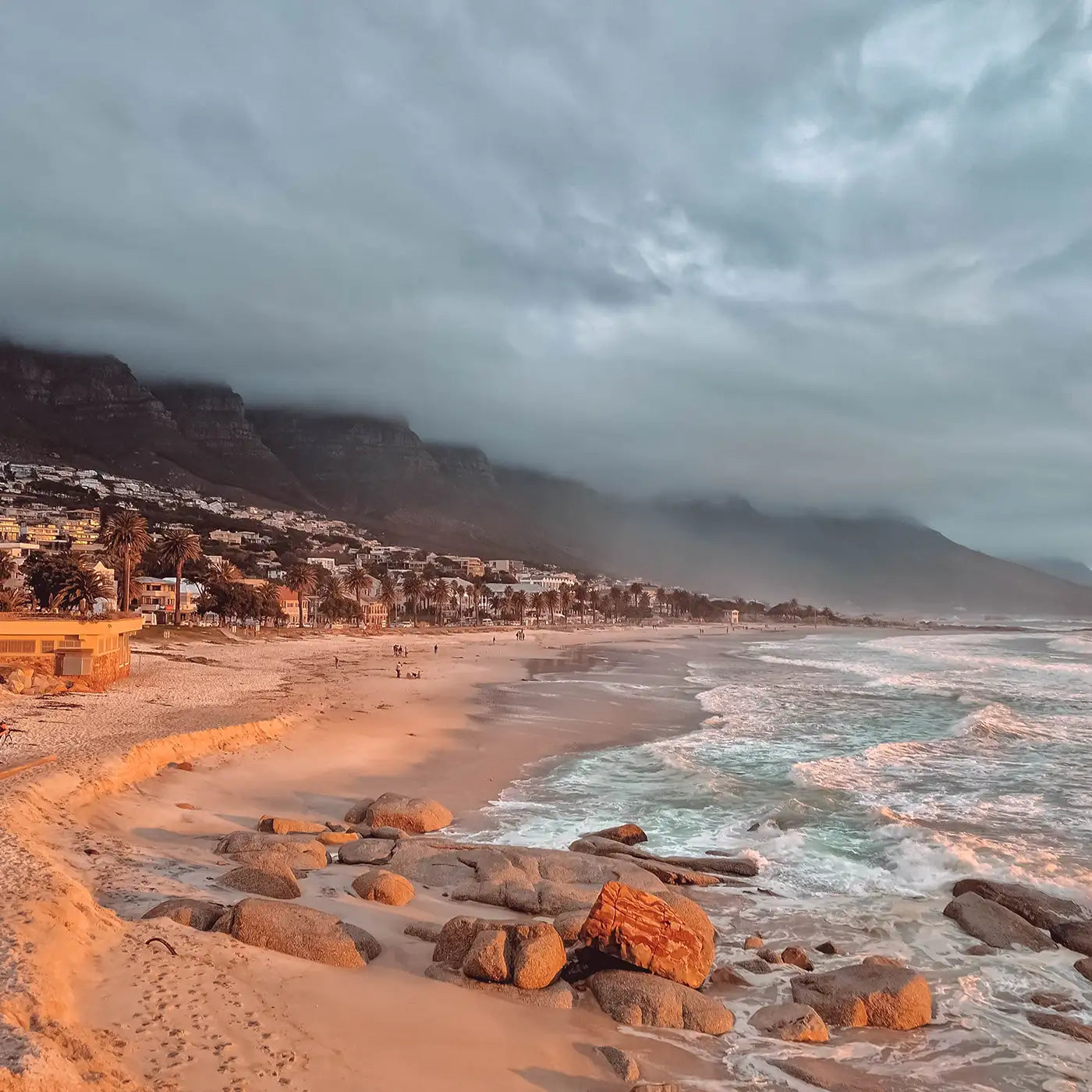 Sandy beach with rocky shore at sunset, mountains shrouded in clouds in the background. Fabulous Flowers and Gifts. Cape Town.