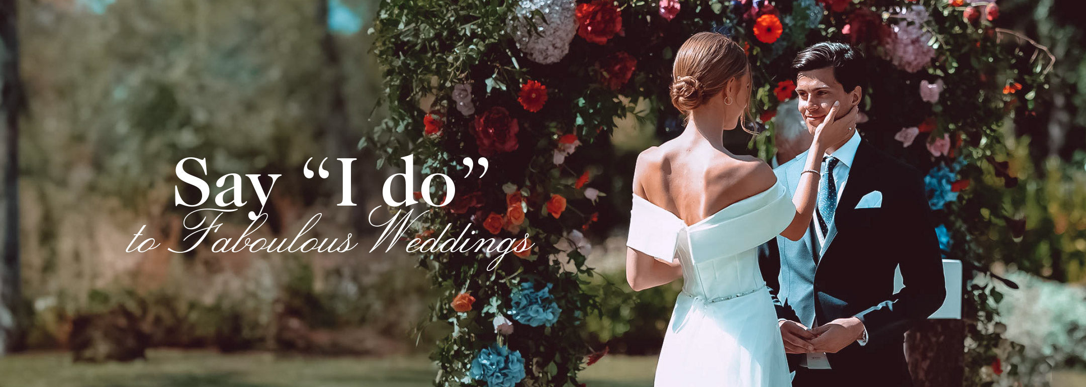 Bride in an off-shoulder white dress tenderly touching groom's face, standing under a colourful flower arch with text 'Say "I do" to Fabulous Weddings.' Weddings at Fabulous Flowers and Gifts.
