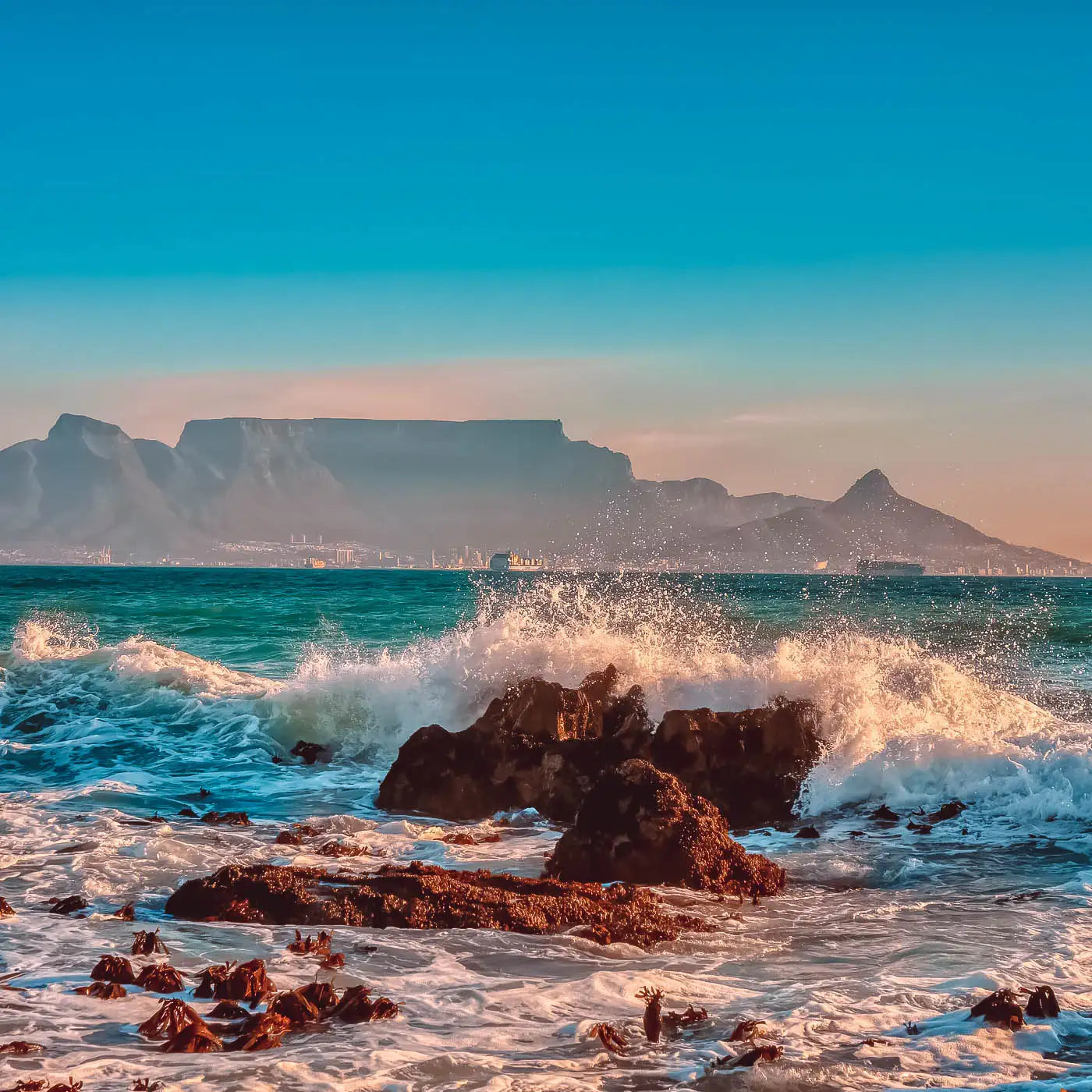 Waves crashing against rocks with Table Mountain in the background under a clear blue sky. Fabulous Flowers and Gifts. Cape Town.