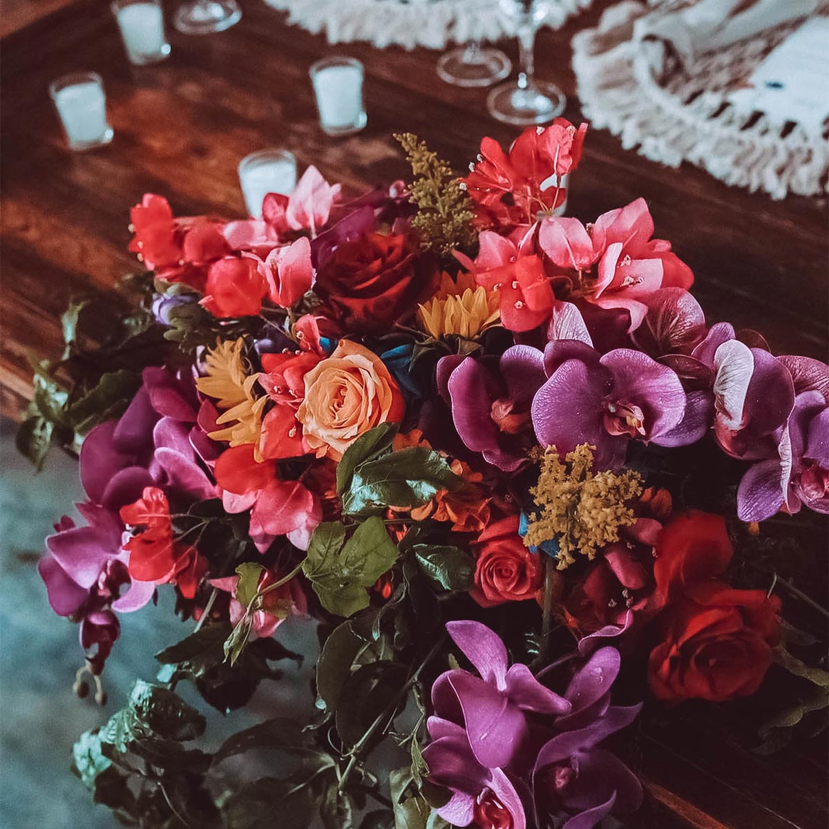 Vibrant floral arrangement with purple orchids, red and pink roses, and greenery on a wooden table set with glasses and candles. Weddings at Fabulous Flowers and Gifts.