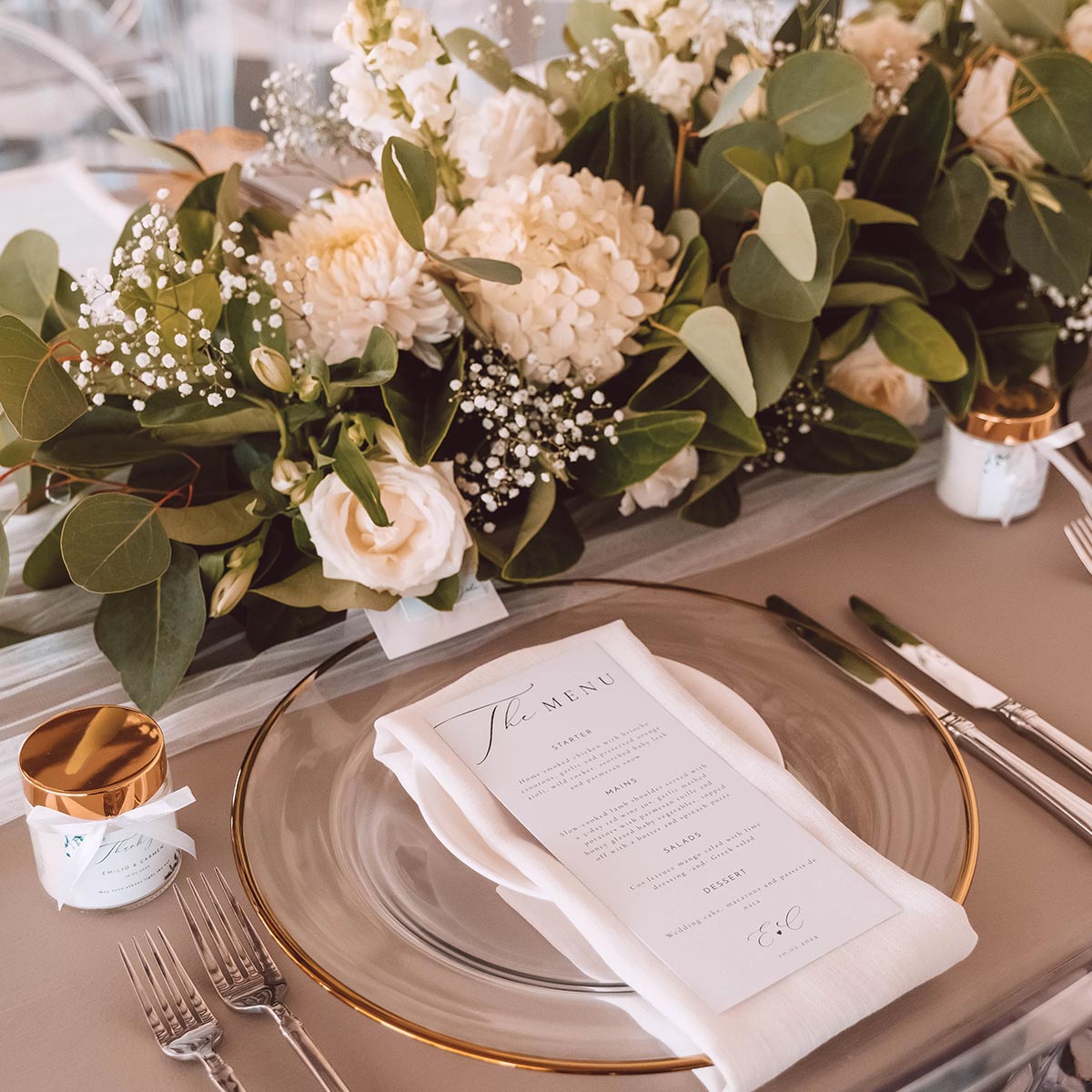 Elegant wedding table setting featuring a clear glass plate with a white napkin and menu, surrounded by lush white floral arrangements and greenery. Fabulous Flowers and Gifts. Weddings.