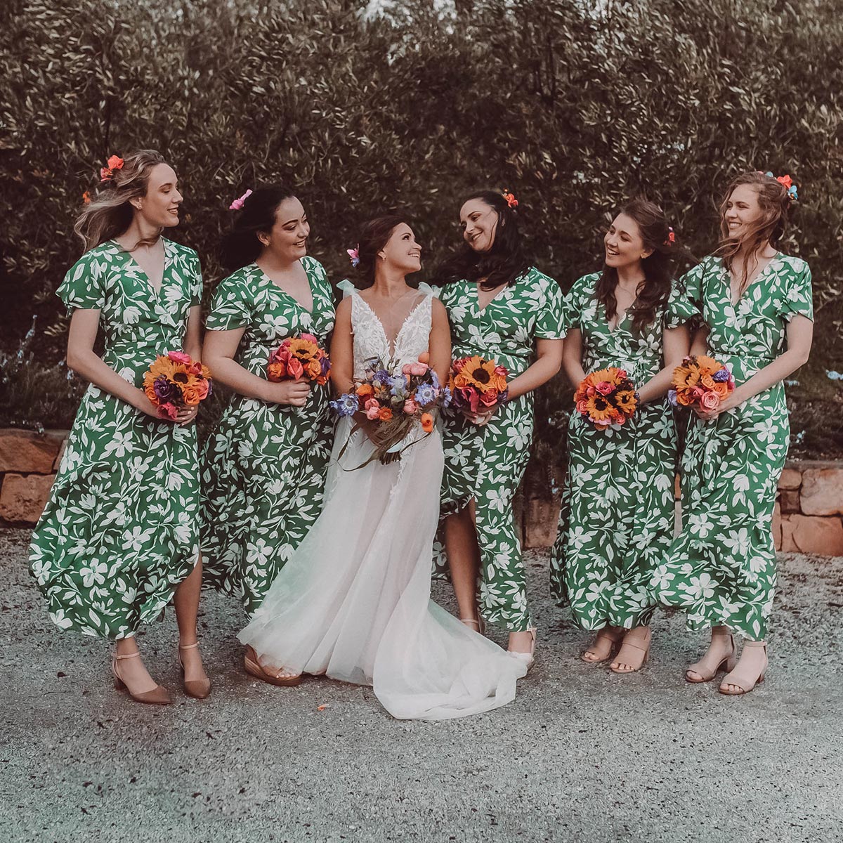 Bride in white gown with bridesmaids in green floral dresses holding colourful bouquets, celebrating outdoors. Fabulous Flowers and Gifts. Weddings