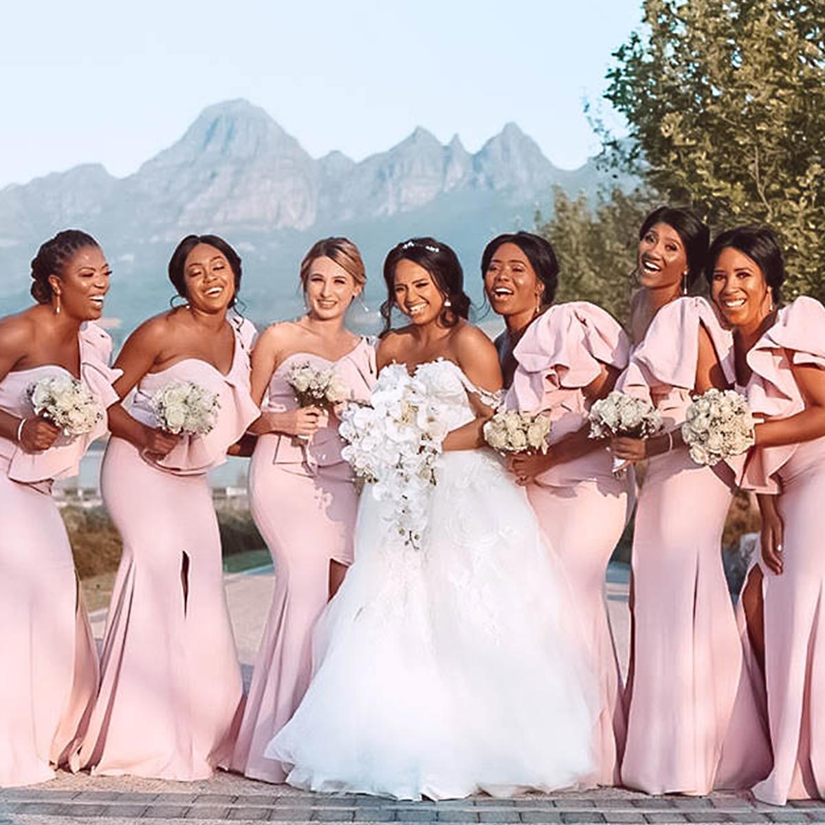 Bride with bridesmaids in matching blush pink dresses holding elegant bouquets against a mountain backdrop, showcasing wedding floral designs. Fabulous Flowers and Gifts.