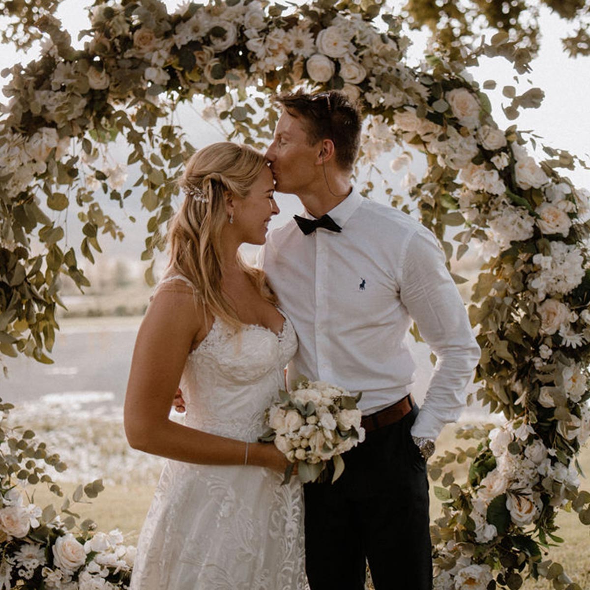 Bride and groom share a tender moment under a lush floral arch adorned with white roses and greenery, celebrating their special day. Fabulous Flowers and Gifts. Weddings.