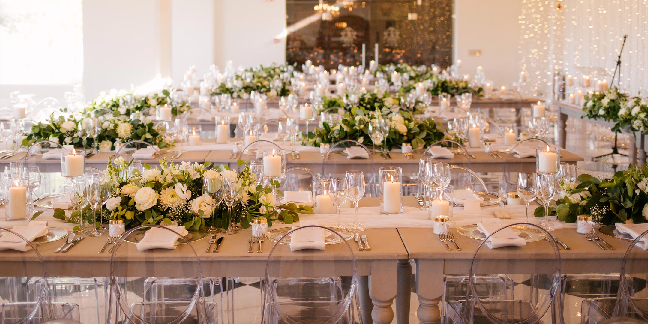 Elegant wedding reception setup with long tables adorned with lush white floral centrepieces, candles, and fine glassware. Fabulous Flowers and Gifts. Weddings.